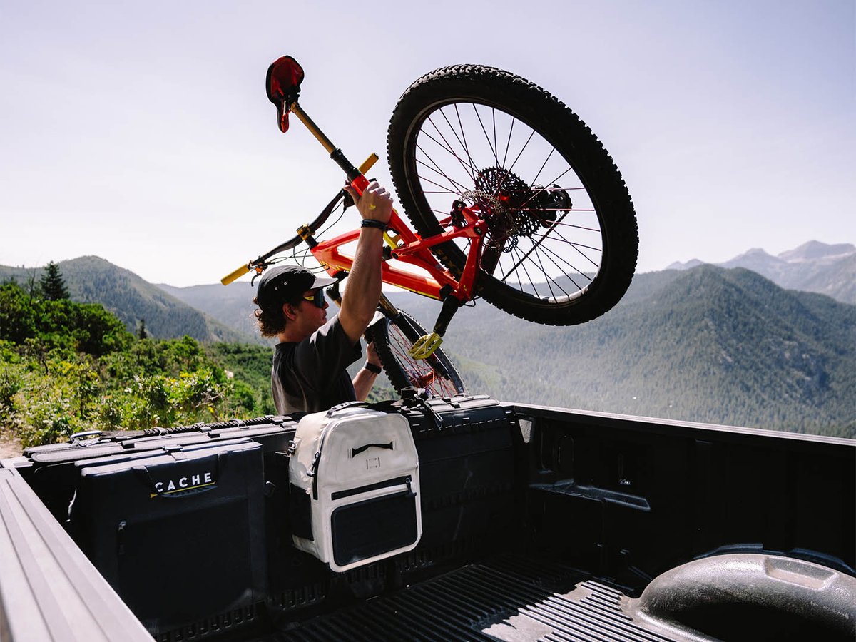 Rider lifting an orange full-suspension mountain bike into a truck bed fitted with a black CACHE tailgate pad, green peaks behind.