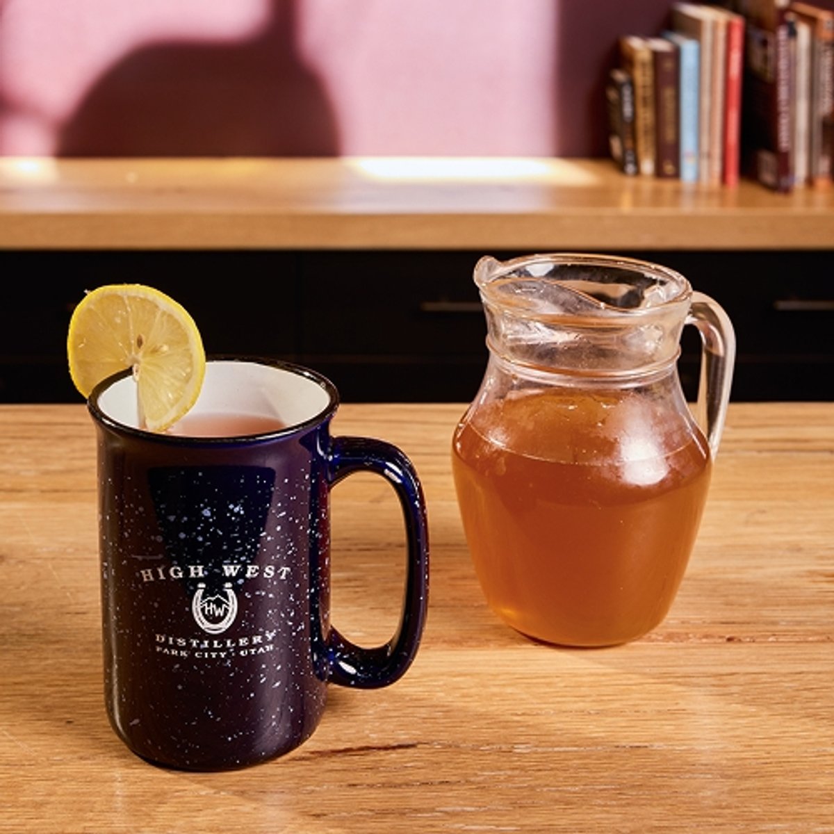A bar countertop displays a mug with a lemon wheel, a glass pitcher of honey syrup, a bottle of High West Double Rye, and a glass jug ginger juice, set against shelves filled with liquor bottles; large text reads "Hot toddy" below the High West Distillery and Backcountry logos.