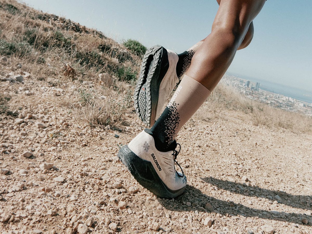 Low-angle view of Salomon Aero Blaze 4 GRVL GTX on rocky gravel terrain, outsole and upper visible, city skyline in background