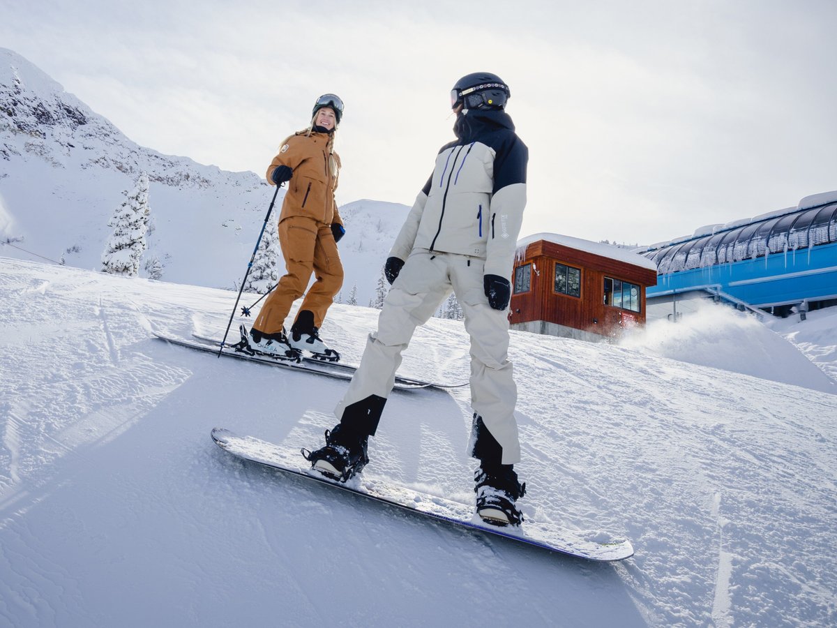 A skier and a snowboarder stand at the top of a groomed run near the top of the chairlift.