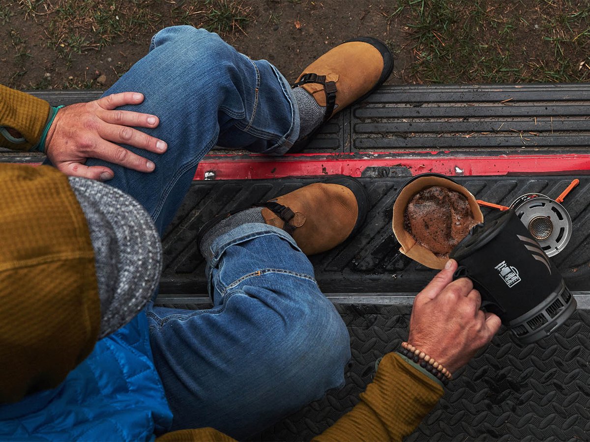 Overhead view of a person sitting on a truck tailgate in tan Bedrock Mountain Clogs and jeans, making coffee on a camp stove