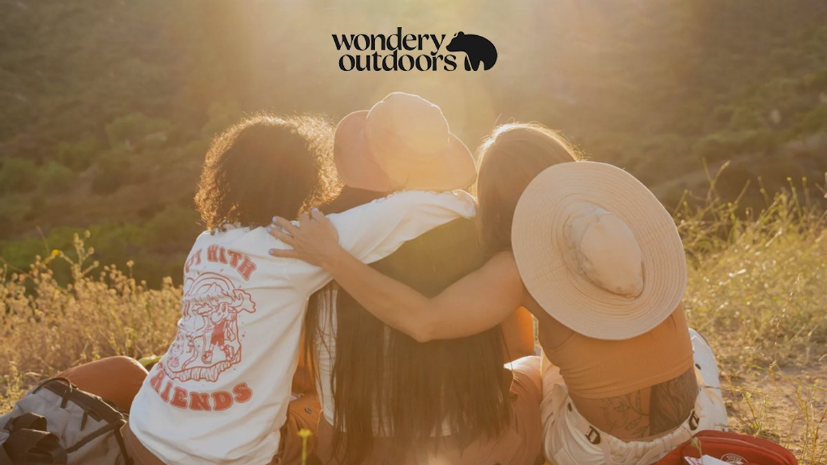 Three friends wearing wide-brimmed sun hats embrace during a golden hour outdoor adventure in a scenic natural landscape.