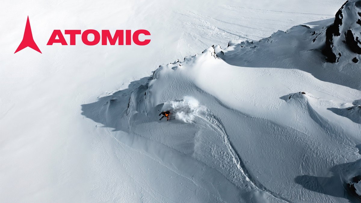 An aerial view shows a lone skier carving a trail through deep powder on a steep, snow-covered mountain ridge under the Atomic logo.