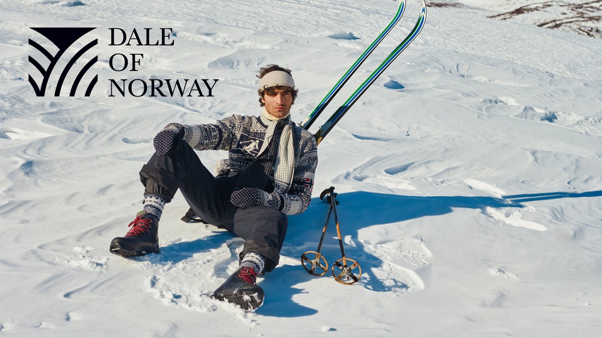 A smiling skier in traditional Norwegian knit sweater and winter gear poses with skis and vintage skates on a snow-covered mountain landscape in Norway.