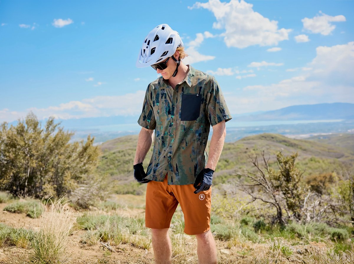 A male mountain biker stands on a high desert ridgeline wearing a patterned olive short-sleeve button-up shirt, rust-orange MTB shorts, black riding gloves, sunglasses, and a white helmet. Rolling hills and a valley vista stretch behind him under a partly cloudy blue sky