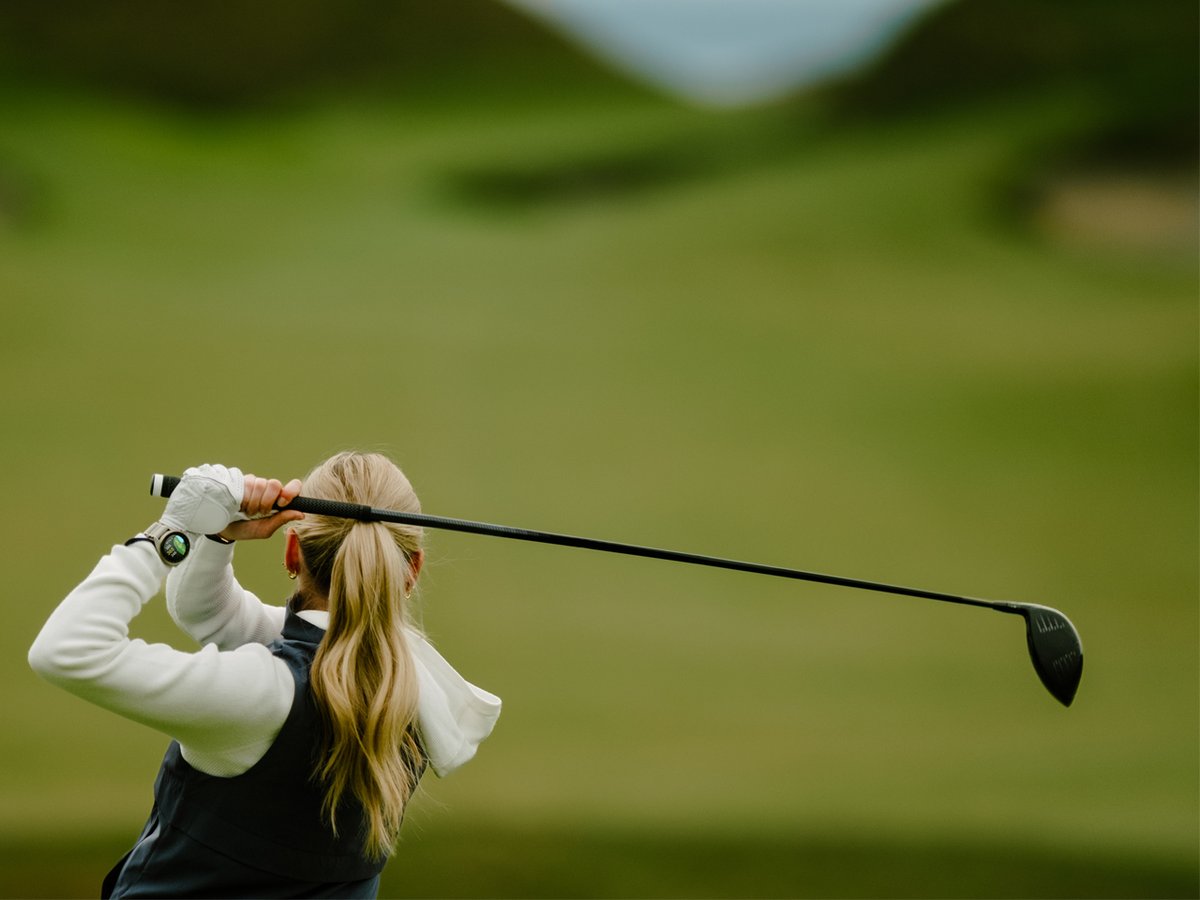 A woman stands in focus, her swung club extended behind her shoulders.