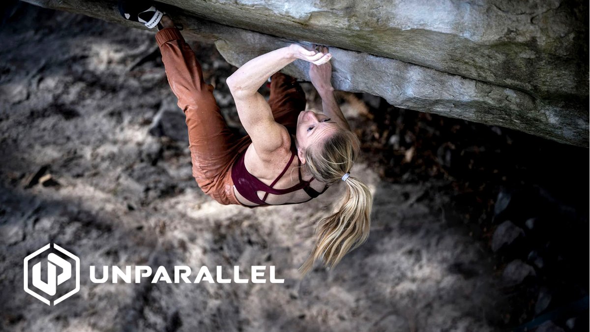 Female rock climber hanging from an overhead boulder using climbing holds while wearing athletic gear and climbing equipment.