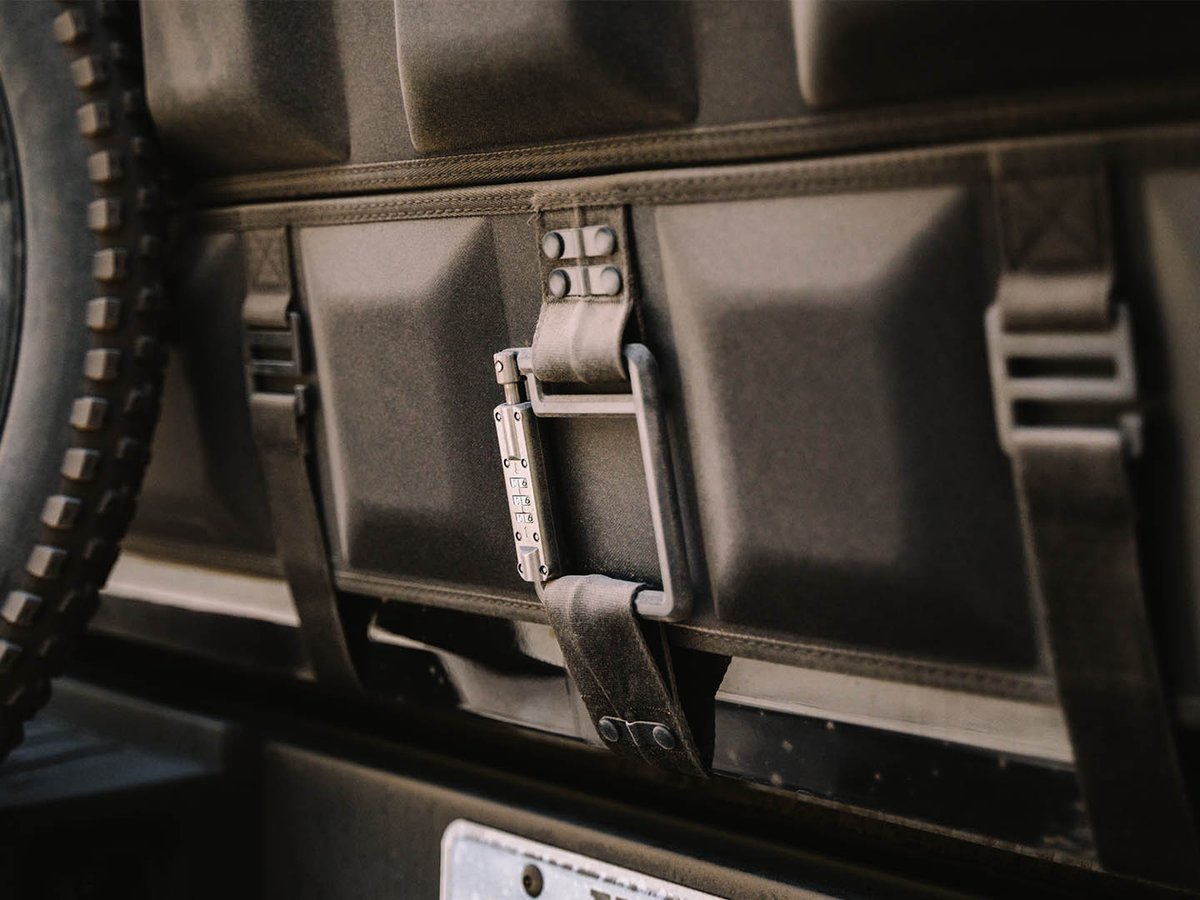 A combination cable lock threaded through a strap on a Cache Basecamp tailgate pad, with a bike tire at the edge of the frame.