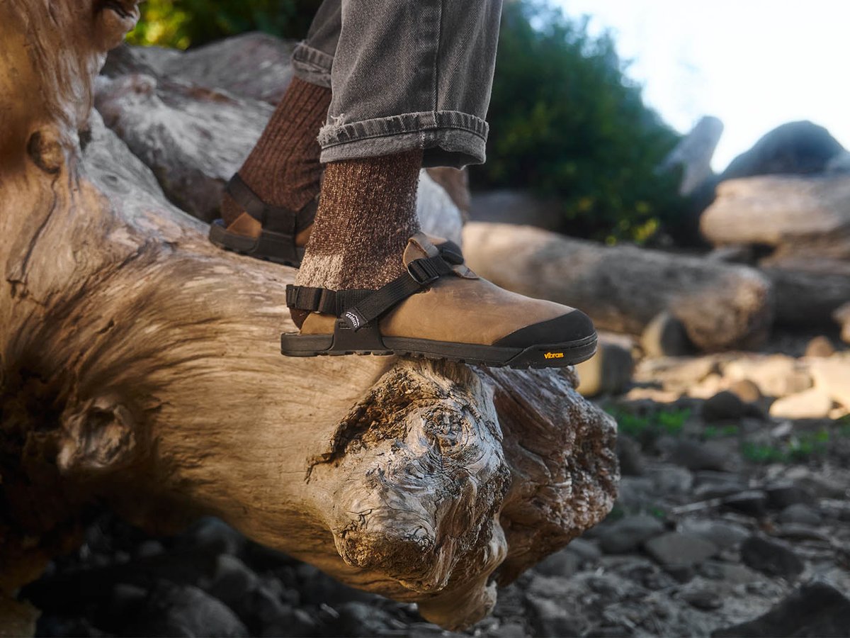 Side profile of Bedrock Mountain Clog in tan nubuck worn with wool socks on driftwood log, showing flat heel-to-toe sole profile and Vibram midsole against rocky beach background