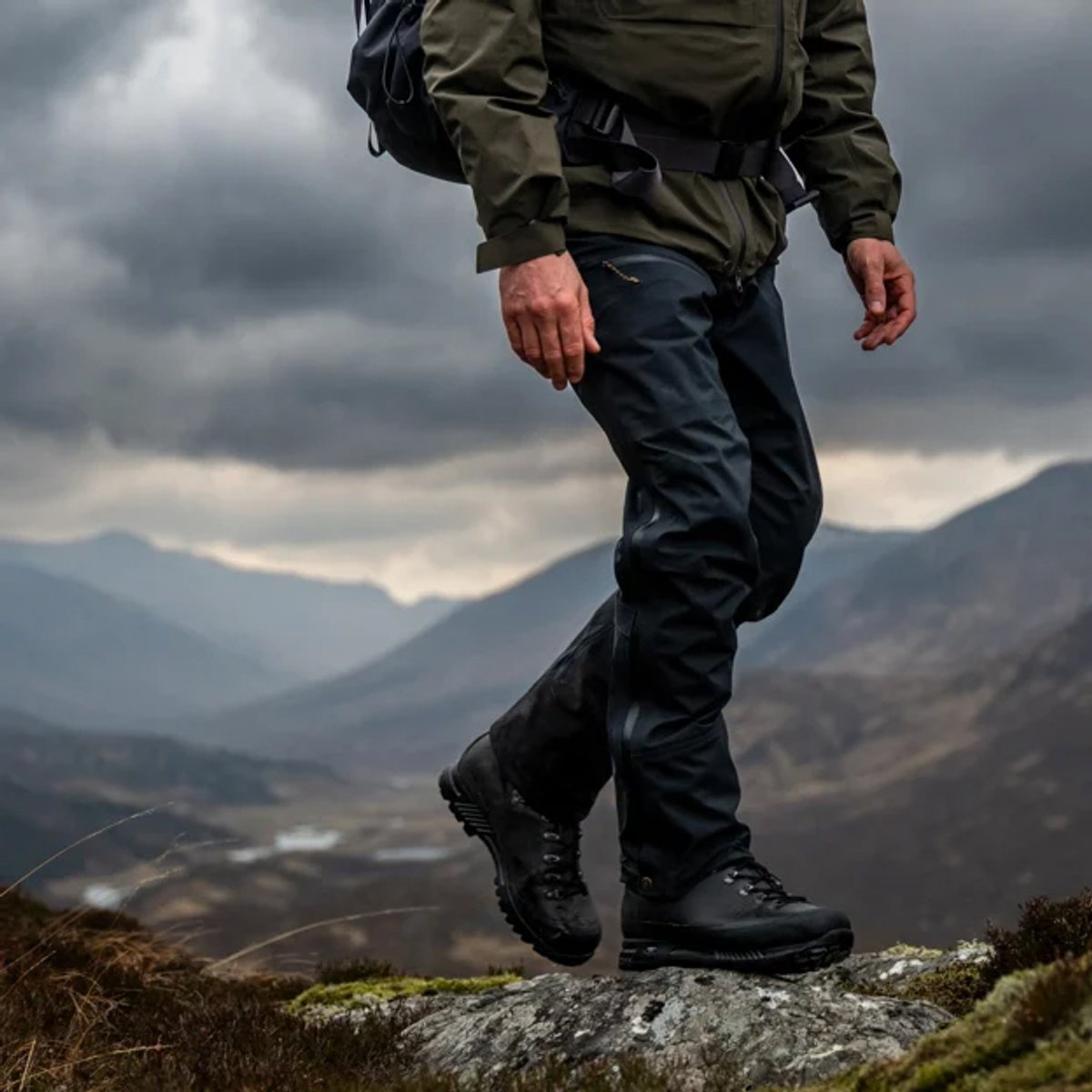 Hiker wearing dark navy hiking pants and olive green jacket standing on rocky mountain terrain with valley landscape and cloudy sky in background.