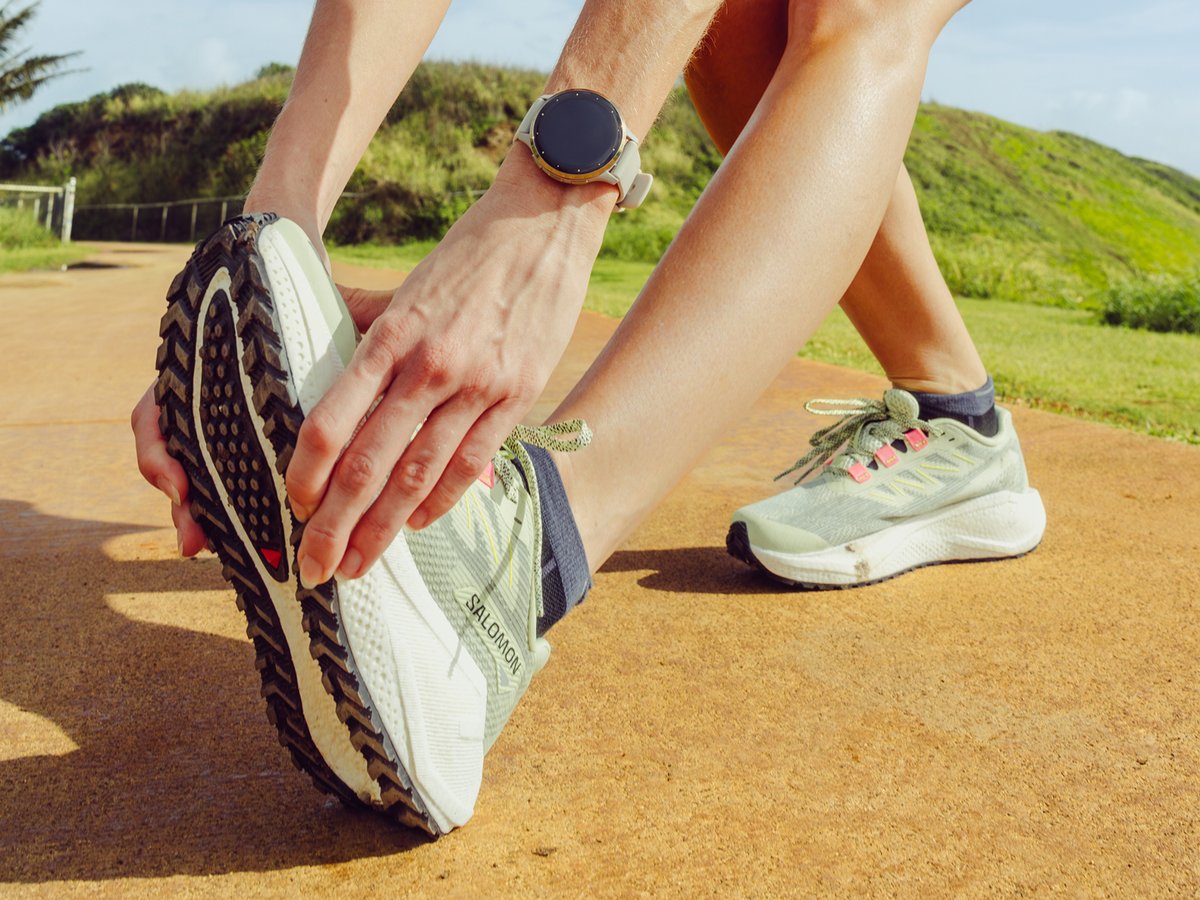 Runner stretching outdoors in Salomon trail shoes with a GPS watch on wrist, against a coastal hillside backdrop.