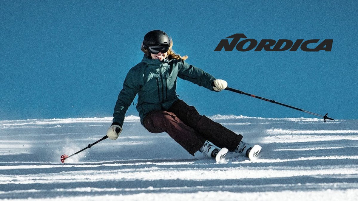 Professional skier in black Nordica gear executing a carving turn on snowy alpine slopes under clear blue sky.