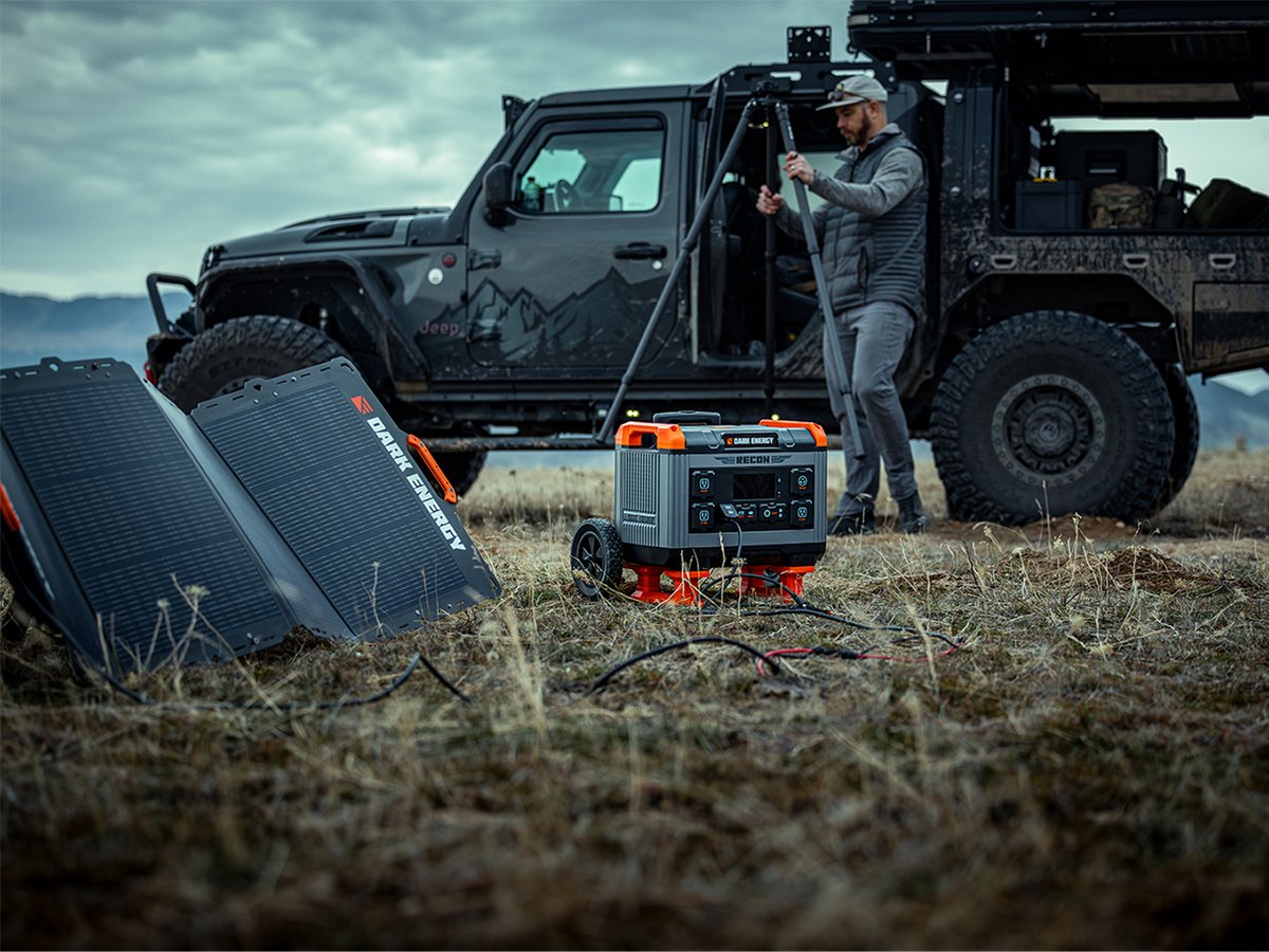 A camper sets up beside a mud-covered Jeep with a rooftop tent while a Dark Energy Recon 2000 power station charges from two Spectre solar panels in the grass.