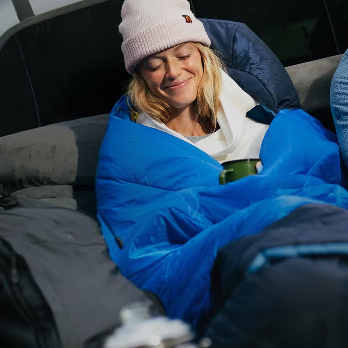 A woman wearing a beanie sits in a car, holding a steaming mug, cozy in her Marmot Wraptor sleeping bag. 