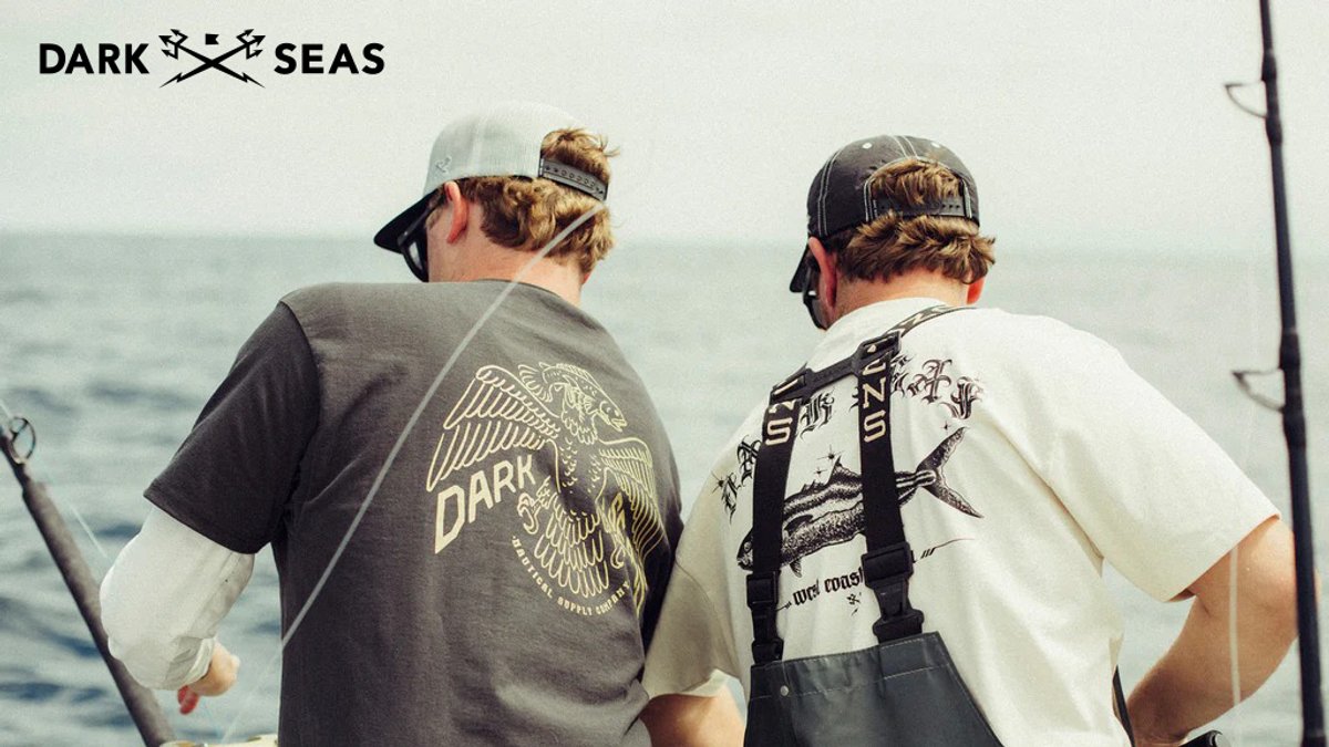 Two anglers wearing Dark Seas branded fishing shirts and caps stand on a boat facing the ocean while holding fishing rods.