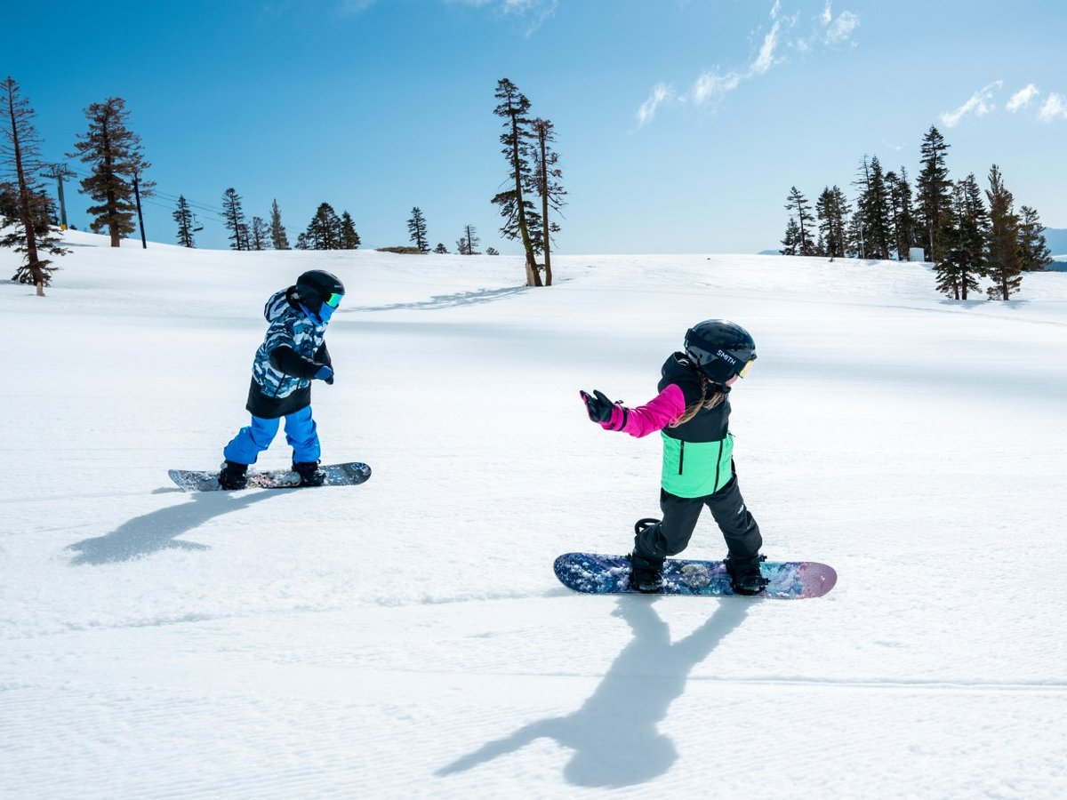 Two young kids snowboarding down a sunny groomed run surrounded by pine trees.