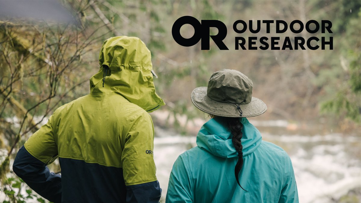 Two people wearing waterproof Outdoor Research rain jackets and a sun hat stand together looking out at a rainy forest landscape.