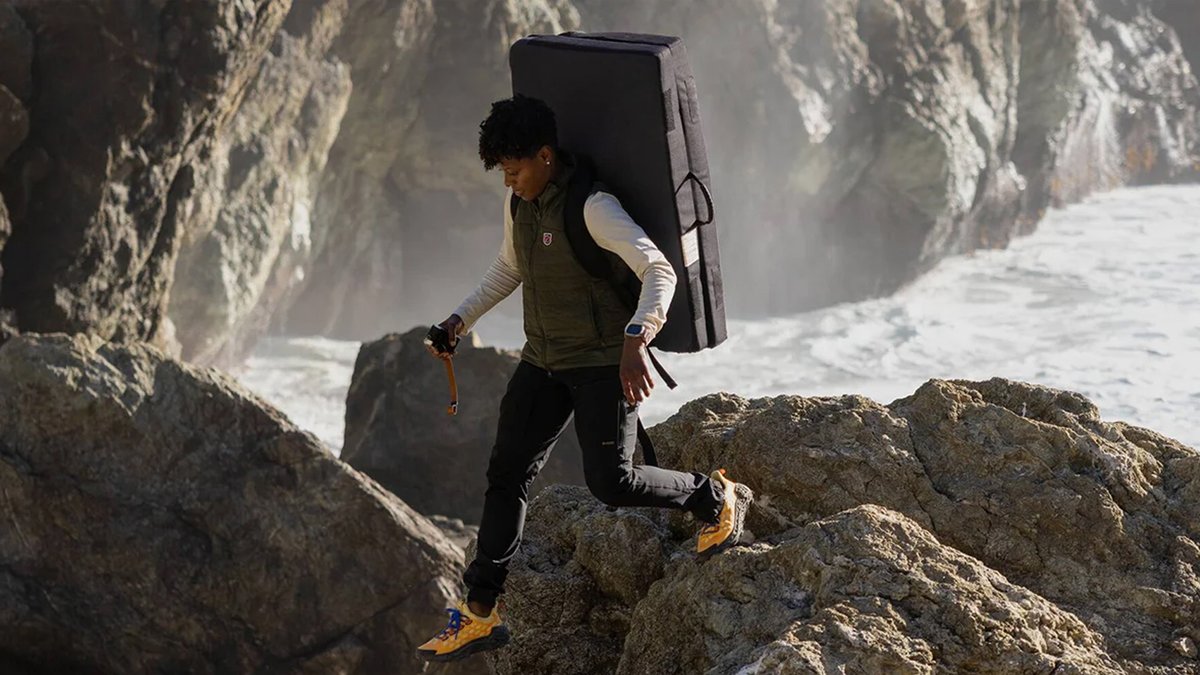 A woman carrying a crash pad walks over rocky borders near a river.