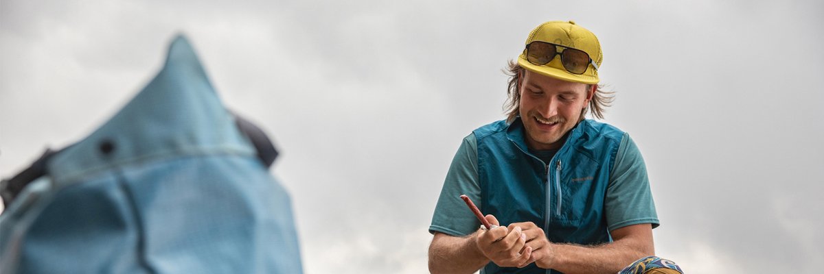 :

Happy male climber in blue shirt and yellow cap inspecting climbing rope with belay device near blue tent outdoors.