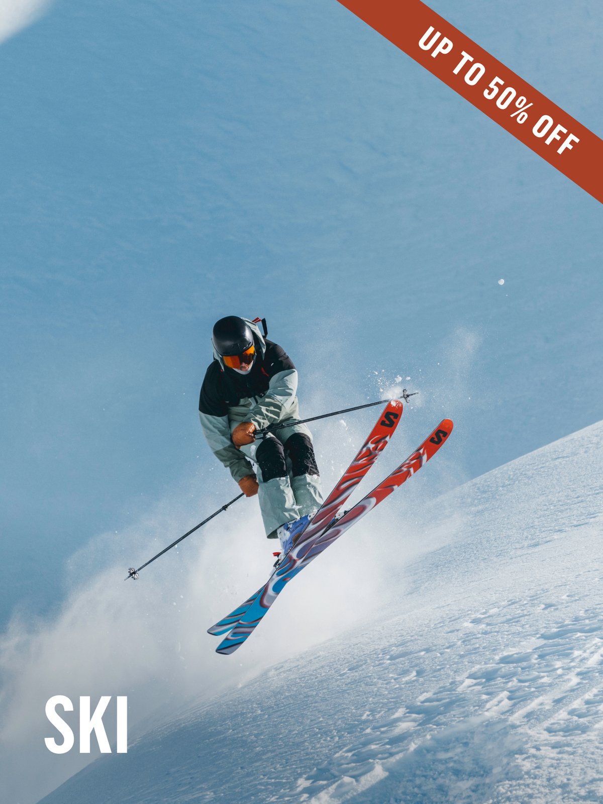 Skier catches air off a steep powder face, Salomon skis crossed mid-jump, helmet and goggles on. Up to 50% off ski gear.