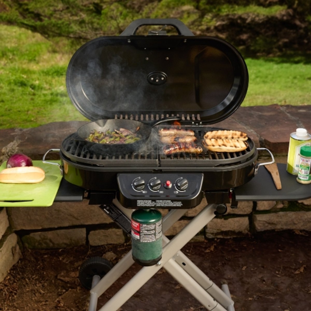 Hot dogs, buns, and a cast iron pan with onions cook on a barbecue camp grill at a campsite. 