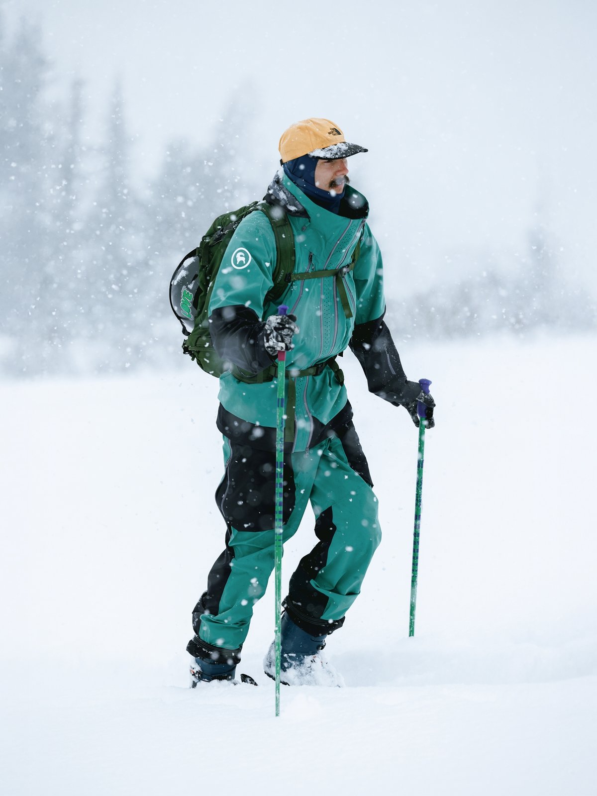 A man on the skintrack on snowy day wearing a green and black jacket and bibs and a tan cap. 