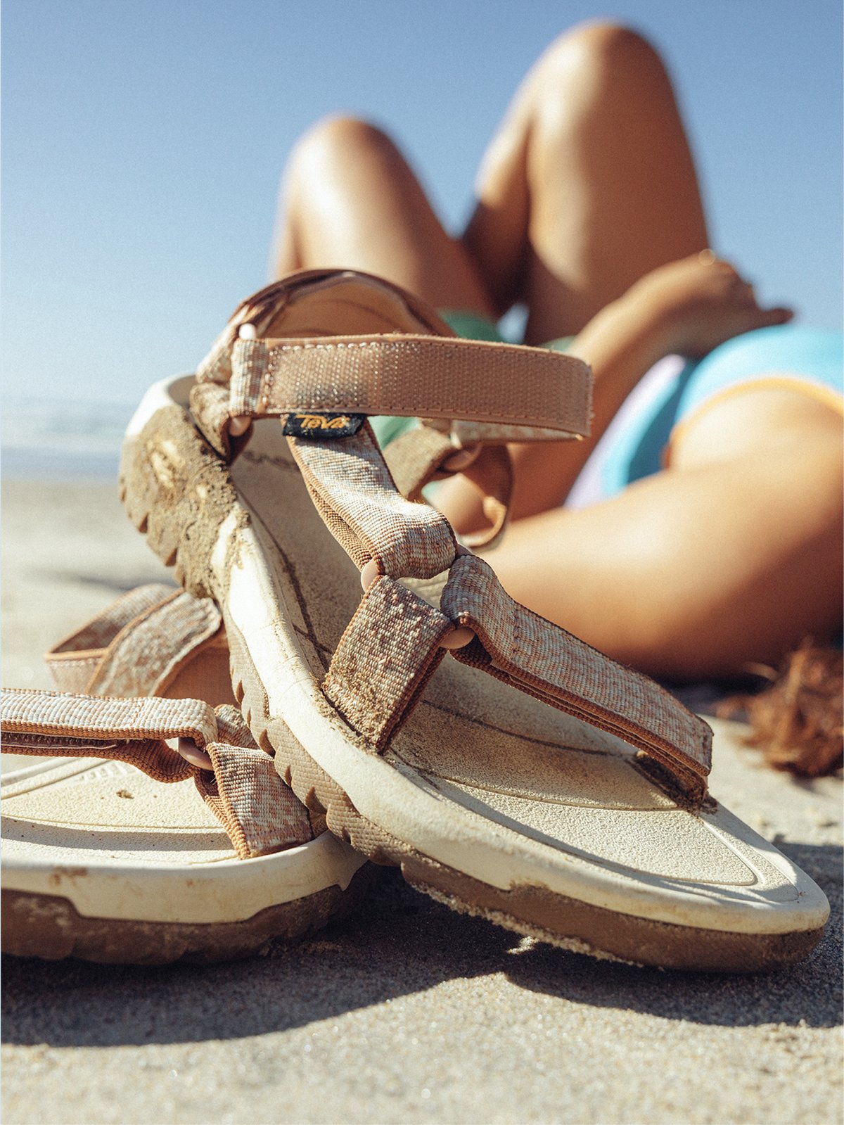 Tan sandals lay on the beach next to a person laying in the sand.