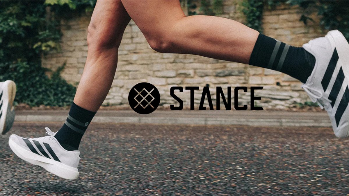 A close-up action shot of a runner's legs in motion on a paved road, wearing black Stance crew socks with grey stripes and white athletic shoes.