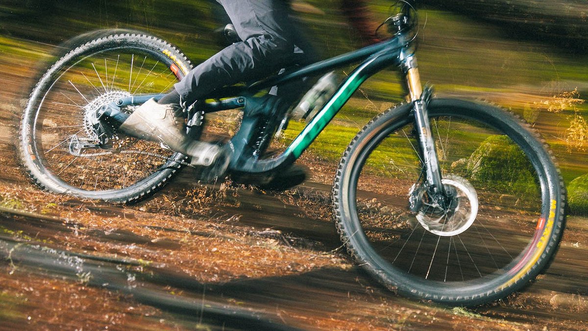 Panned motion shot of a teal full-suspension mountain bike on a trail, showing Reserve aluminum wheels with yellow-sidewall tires and heavy motion blur.