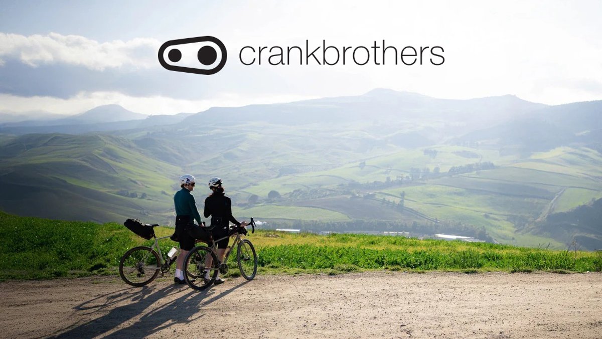 Two cyclists in helmets stand with their road bikes on a hilltop overlooking a sprawling green valley and distant mountains on a clear day.