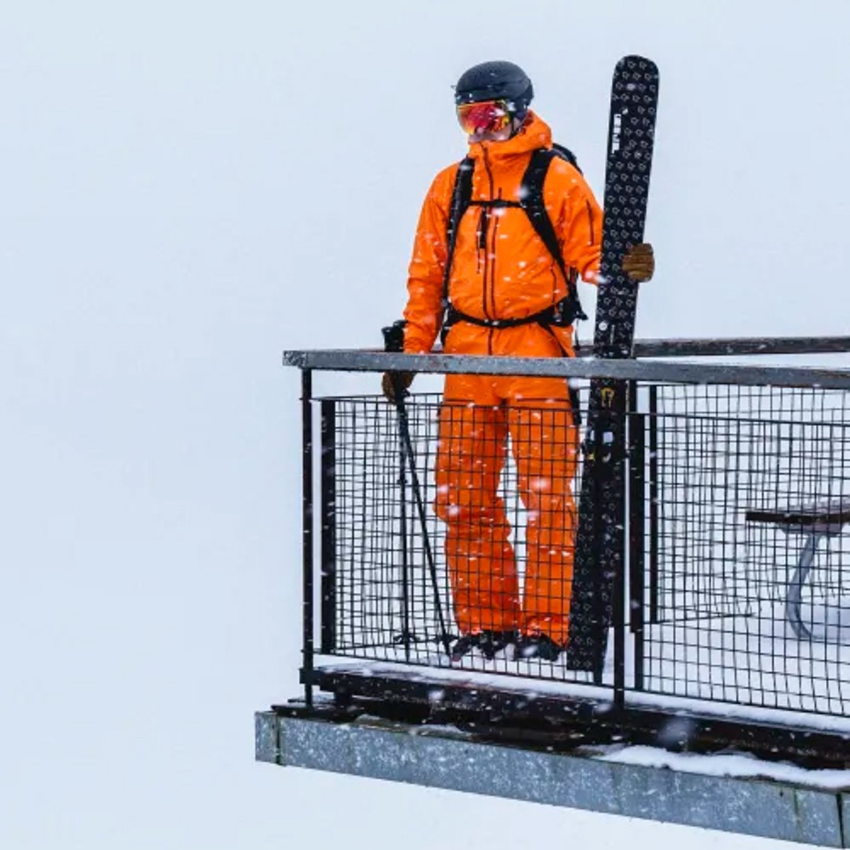 Skier in bright orange winter jacket and helmet standing on a mountain lift platform holding skis in snowy conditions.