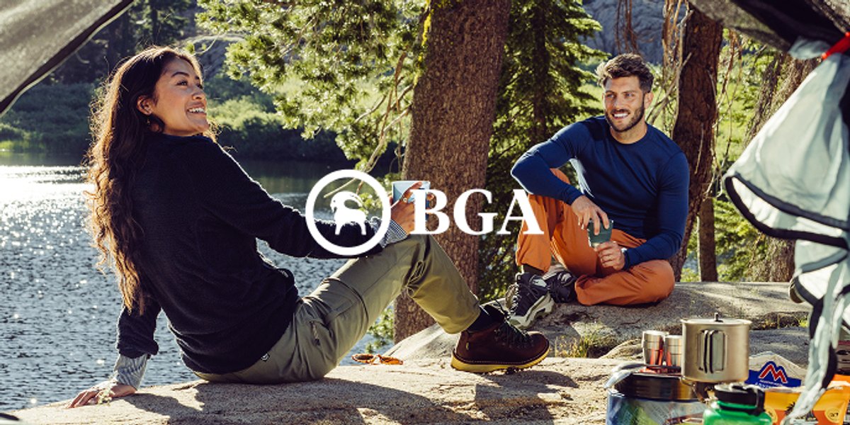 Five friends gathered around a small camp stove fire in a pine forest, laughing and sharing whiskey beside a glowing orange tent.