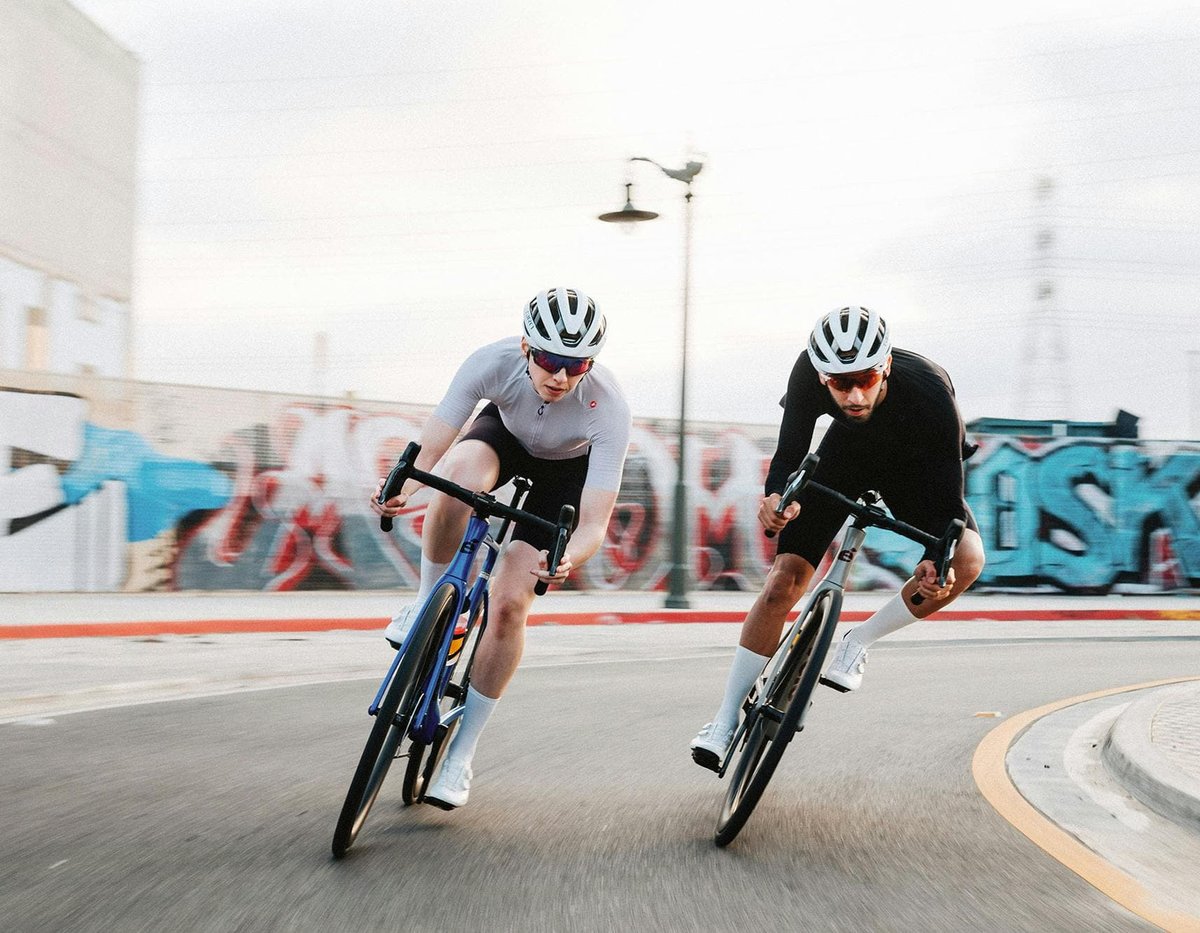 Two road bike riders in aggressive sprint position rounding urban street corner past graffiti wall in city cycling environment