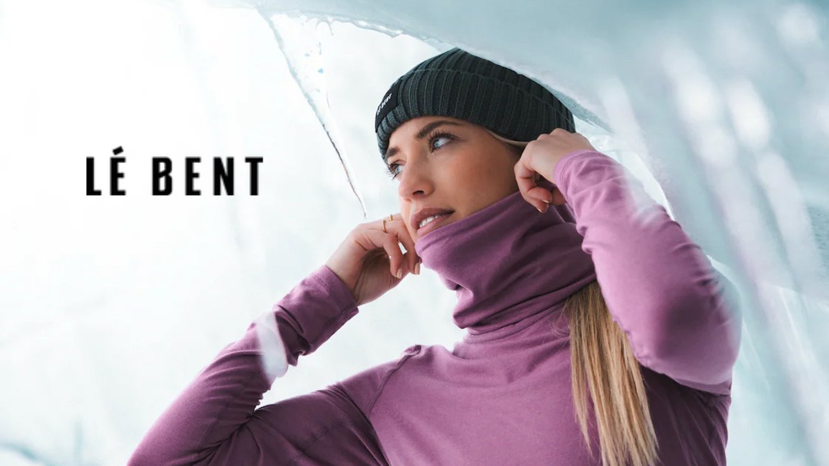 A woman wearing a black beanie and purple jacket stands under an icy outcropping while looking upward against a cloudy sky.