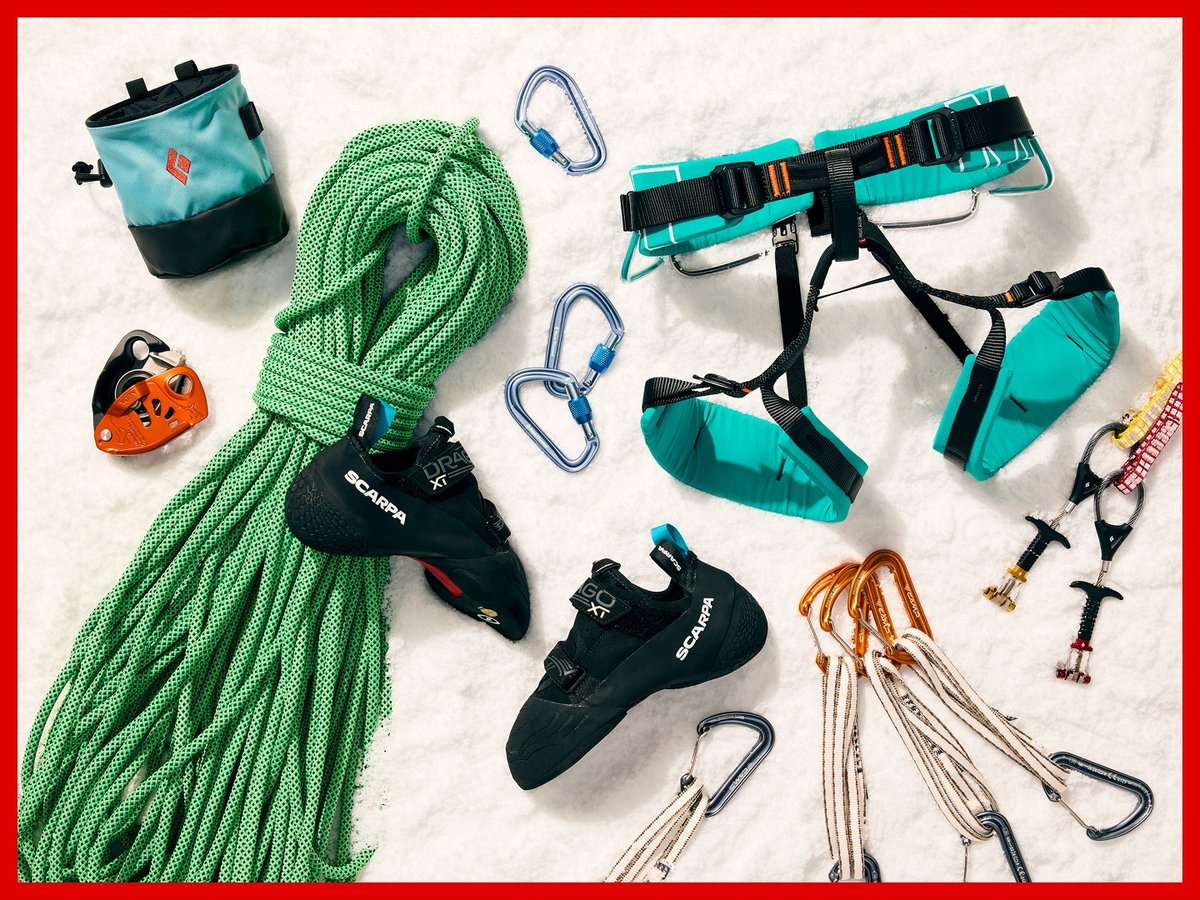 Climbing gear arranged on snow: a green rope, Scarpa climbing shoes, a Petzl Grigri, quickdraws, a harness, and some cams.