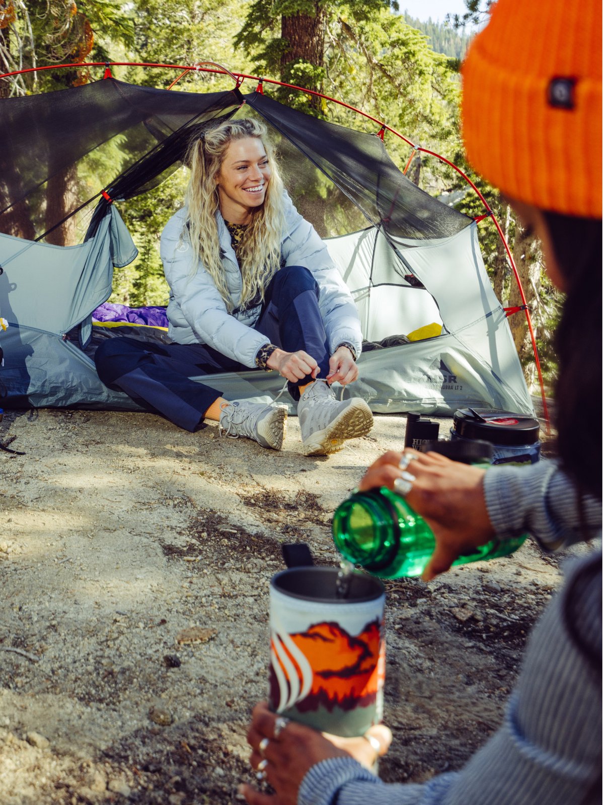 A person with long blonde hair sits at the entrance of a gray camping tent in a forest, tying their hiking boots, while another person in the foreground wearing an orange beanie pours water from a green bottle into a JetBoil.
