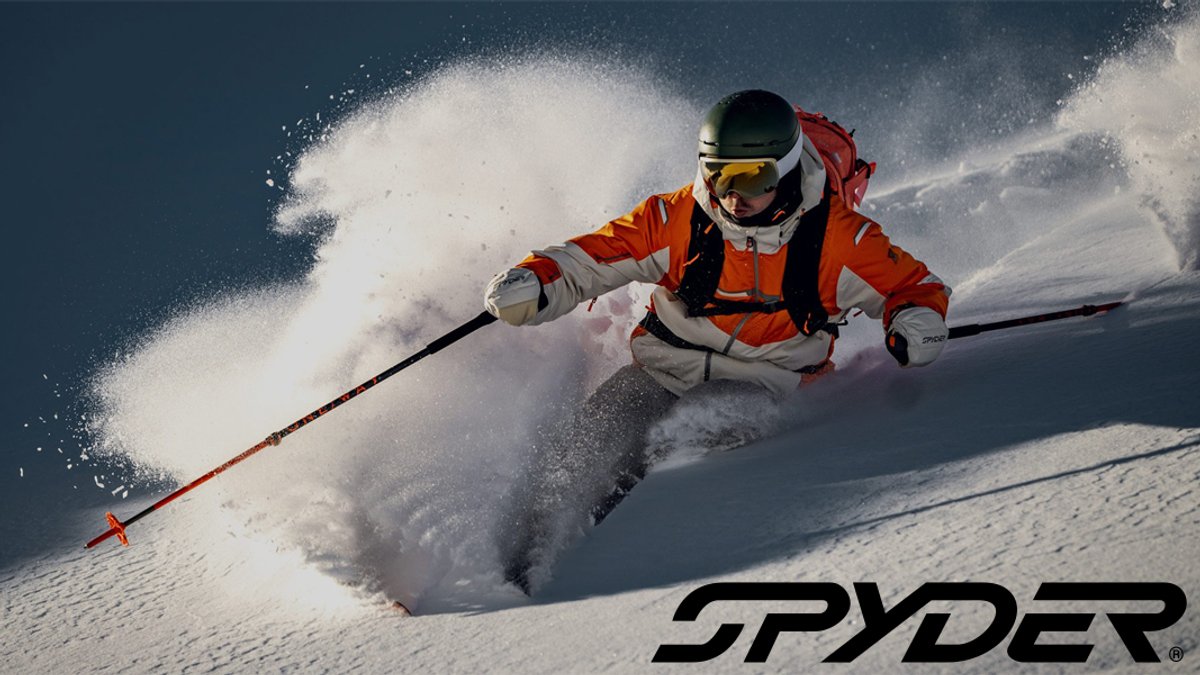 Professional skier in orange jacket and white helmet executing a deep powder turn with Spyder branding visible on snow-covered mountain slope.