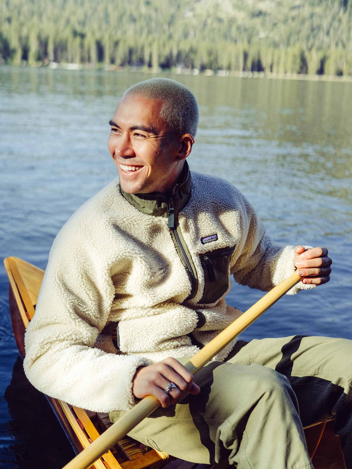 A smiling man wearing a light-colored Patagonia fleece jacket and olive green pants paddles a wooden canoe on a lake.