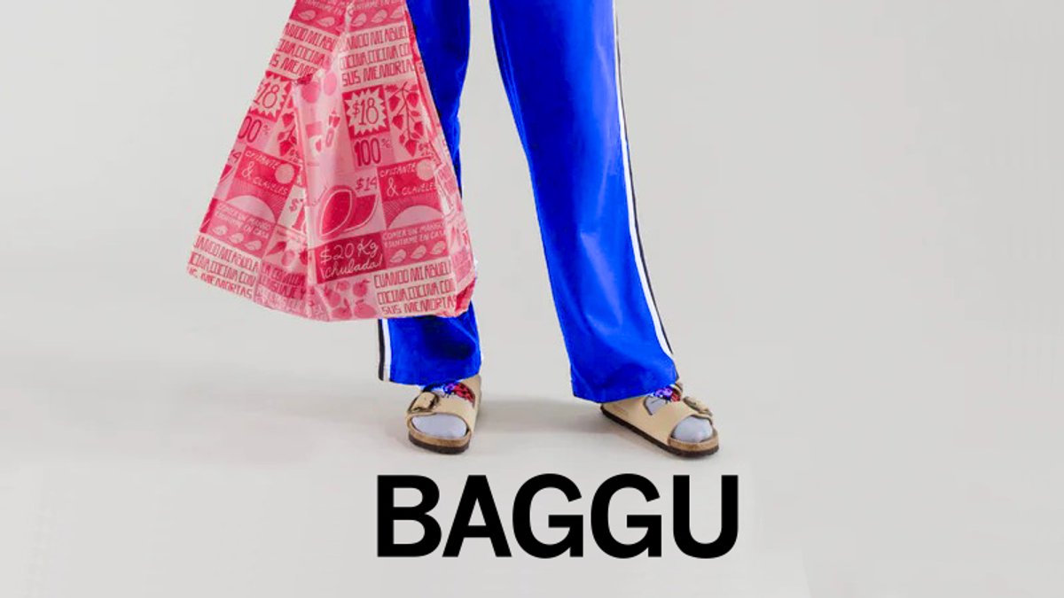 A person wearing bright blue track pants and tan slide sandals holds a pink patterned BAGGU tote against a light background.