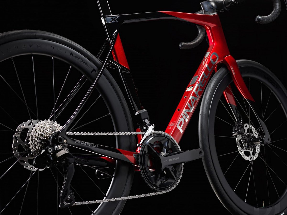 Pinarello road bike red carbon frame with Shimano 105 crankset, cassette, and disc brakes on deep section wheels against dark studio background