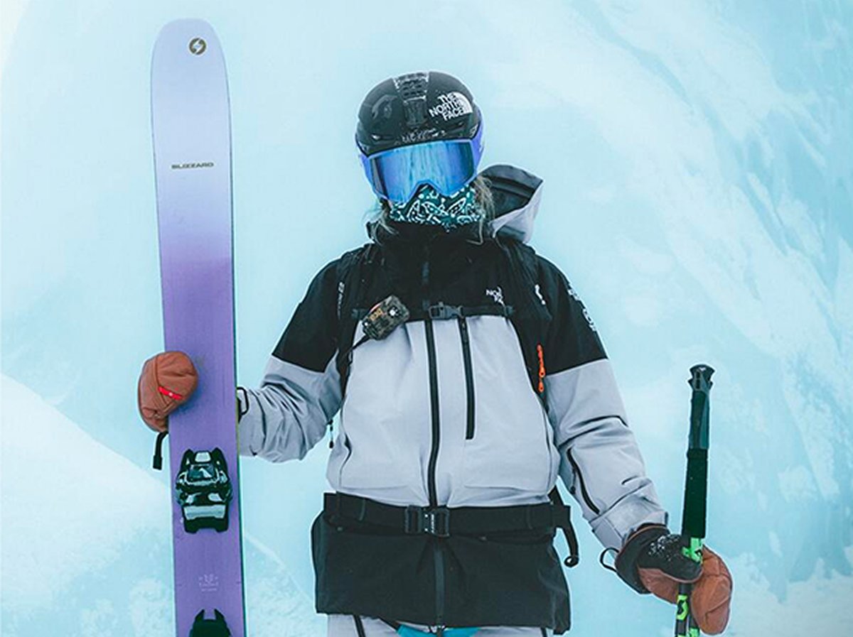 A person in a black helmet and goggles holds a purple ski and a pole while dressed in a winter jacket and gloves.