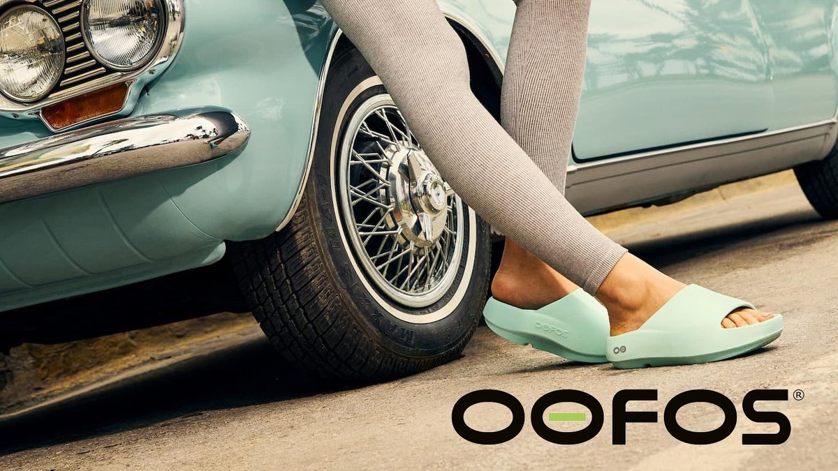 Woman wearing mint green OOFOS slide sandals standing next to a vintage turquoise car with wire wheel rim.