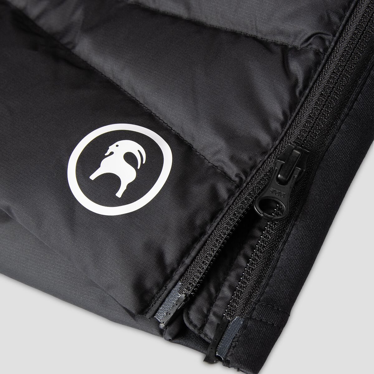 Close-up of the black ripstop shell fabric on a Stansbury ALLIED Down piece, showing the woven texture, reflective goat logo, and zipper detail.