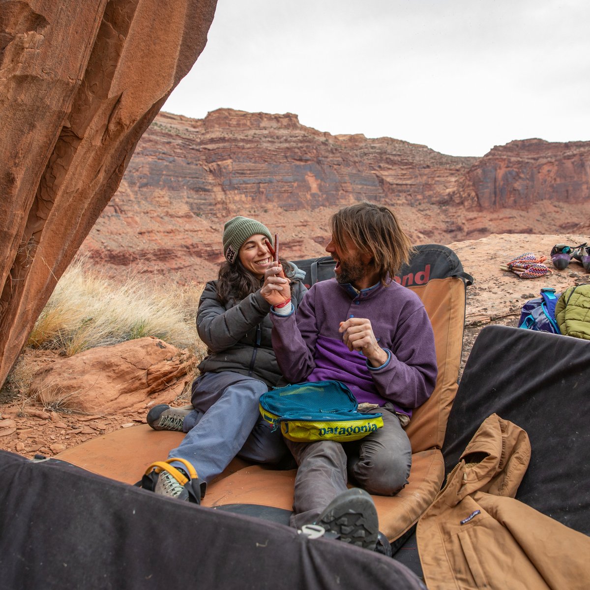 :

Two adventurers enjoy a camping moment under a desert rock shelter wearing outdoor gear while exploring red rock landscapes.