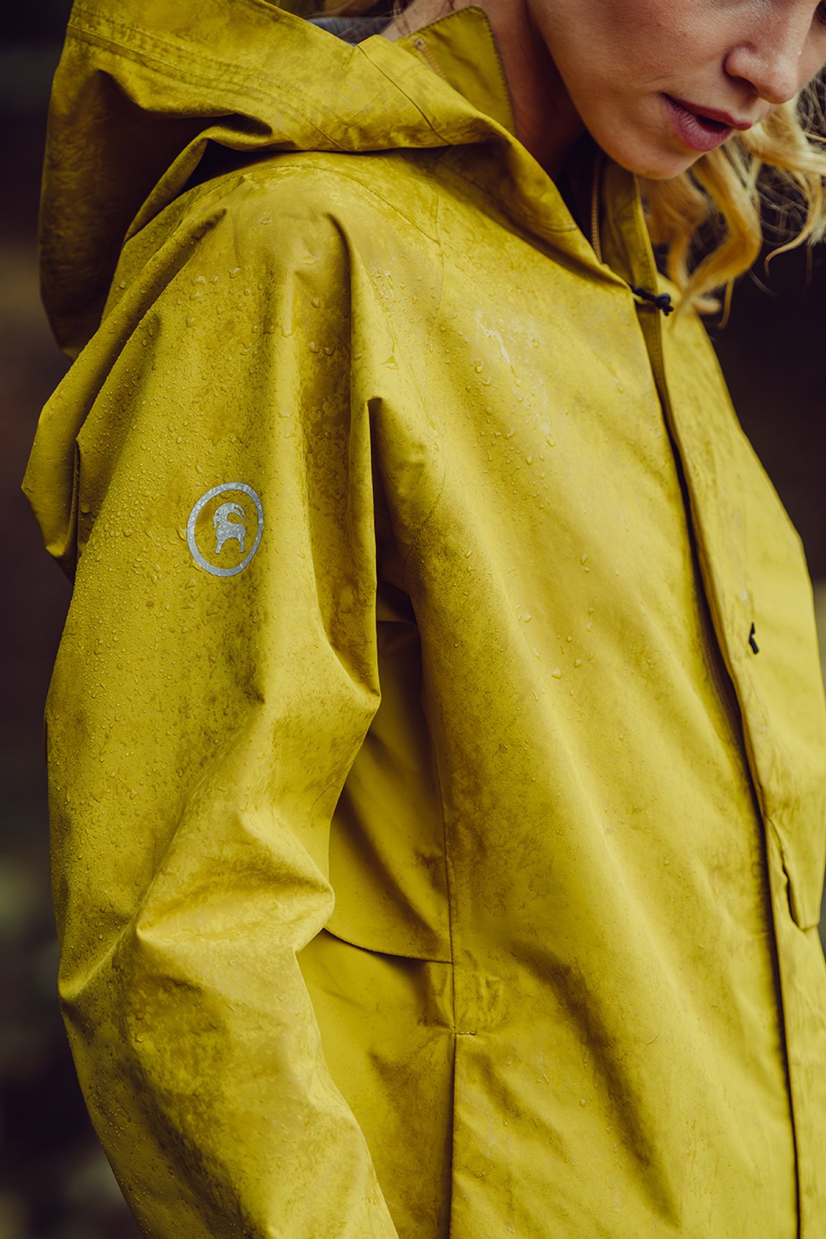 Detail view of wet yellow hooded hardshell jacket showing Backcountry goat logo on sleeve during rainy outdoor conditions