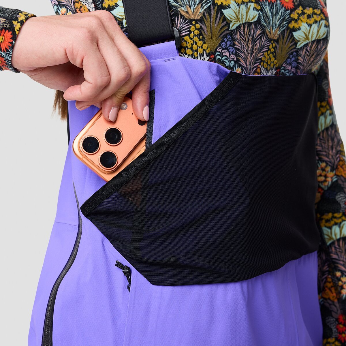 Person pulling phone out of dedicated phone sleeve pocket on upper part of bibs near ribs from below the stretch mesh dump pocket.
