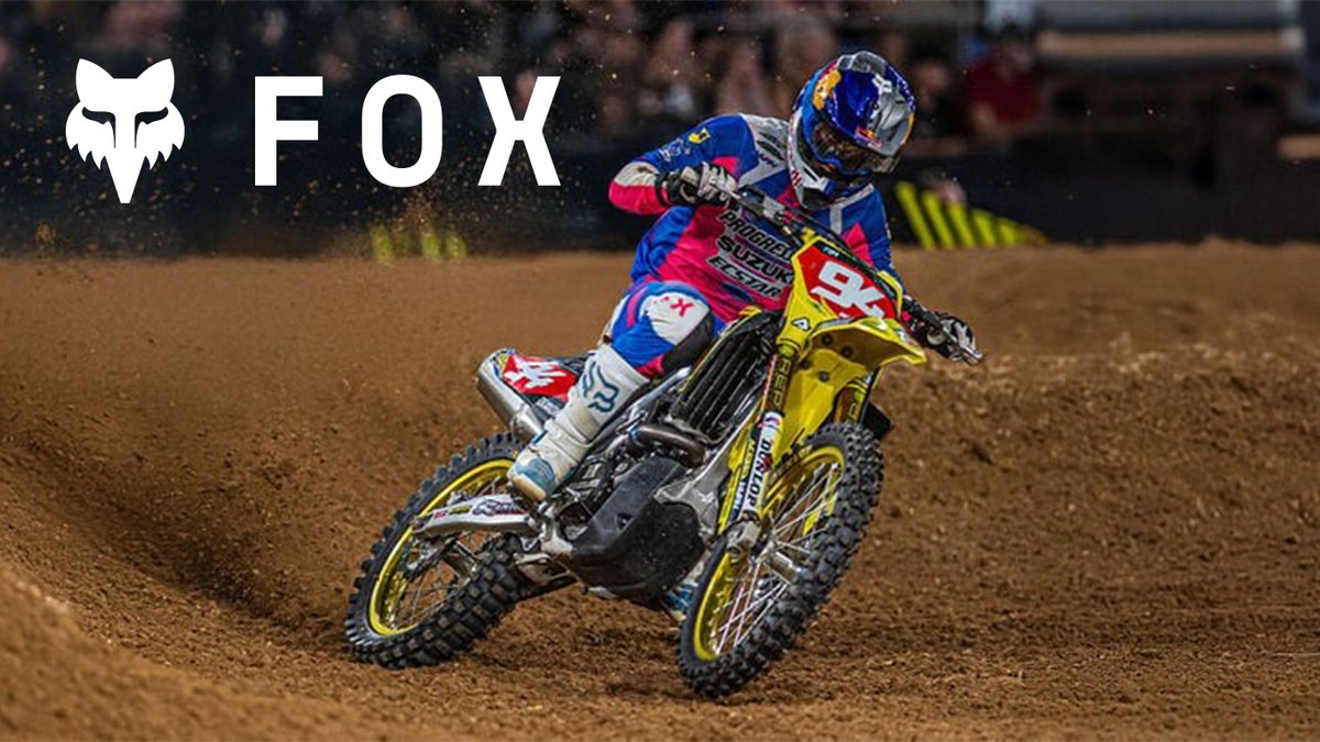 A motocross rider in vibrant blue and pink gear leans into a dirt turn on a yellow racing motorcycle.