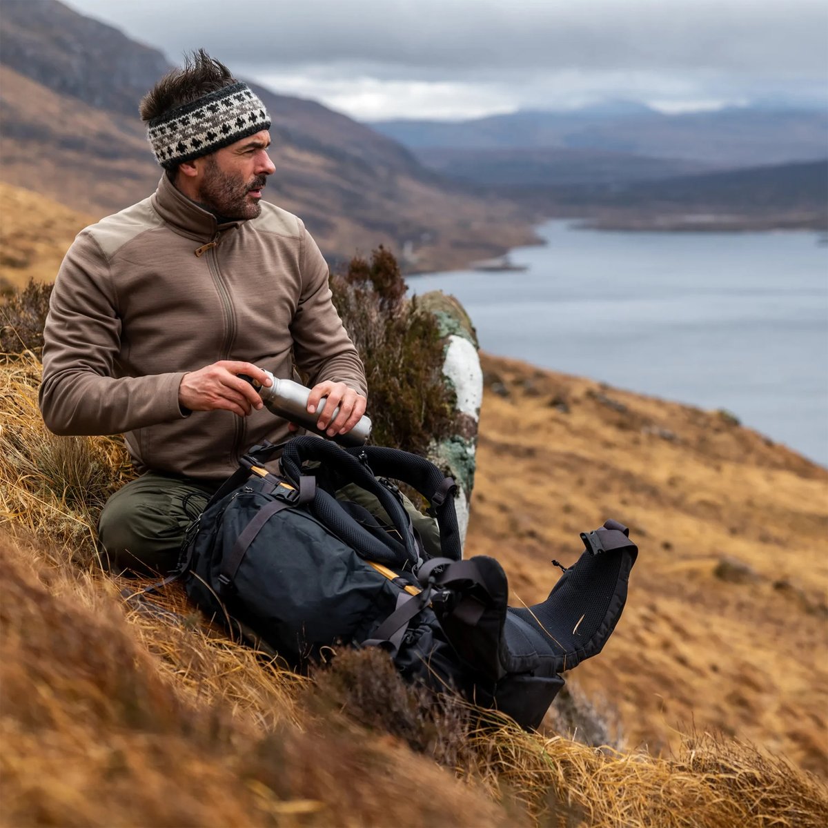 A man sits on a grassy slope, overlooking a lake. He wears a Fjallraven jacket and pants.