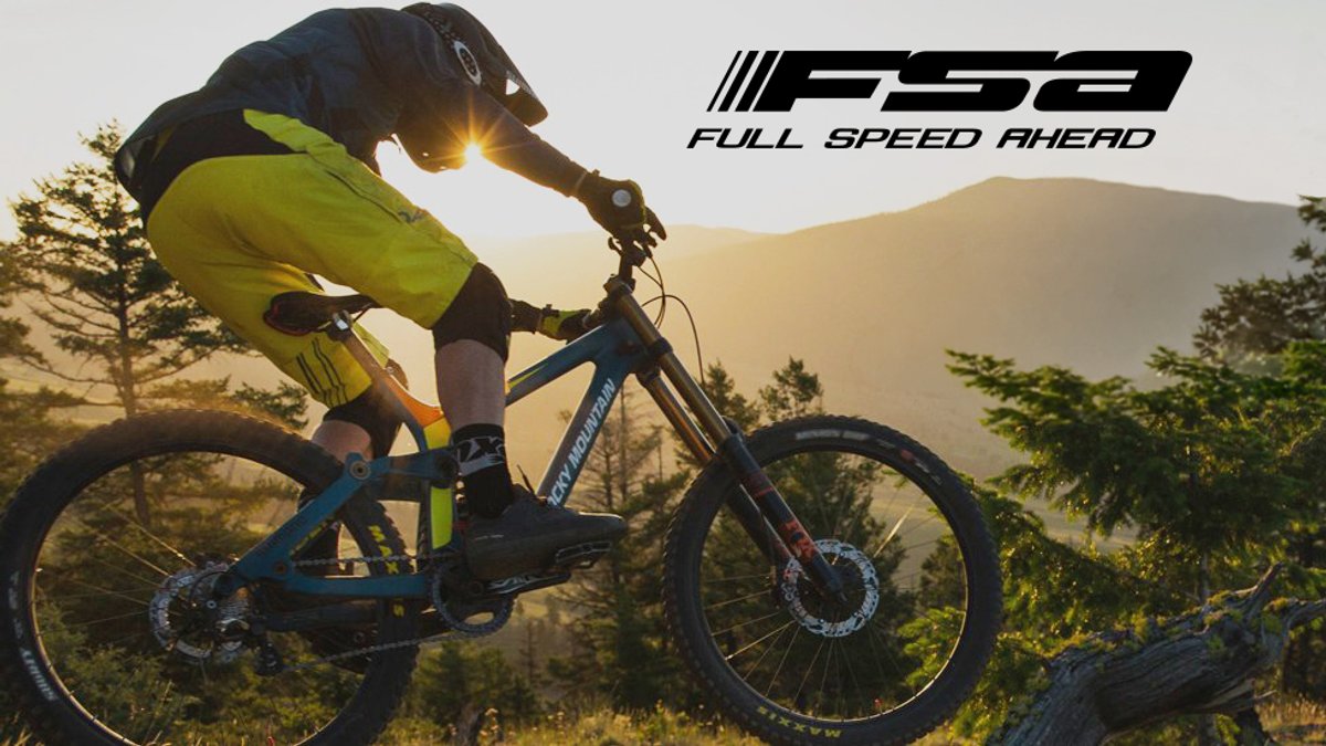 A mountain biker in bright yellow gear rides a blue full-suspension bike down a rocky trail at sunset with forested mountains in the background.