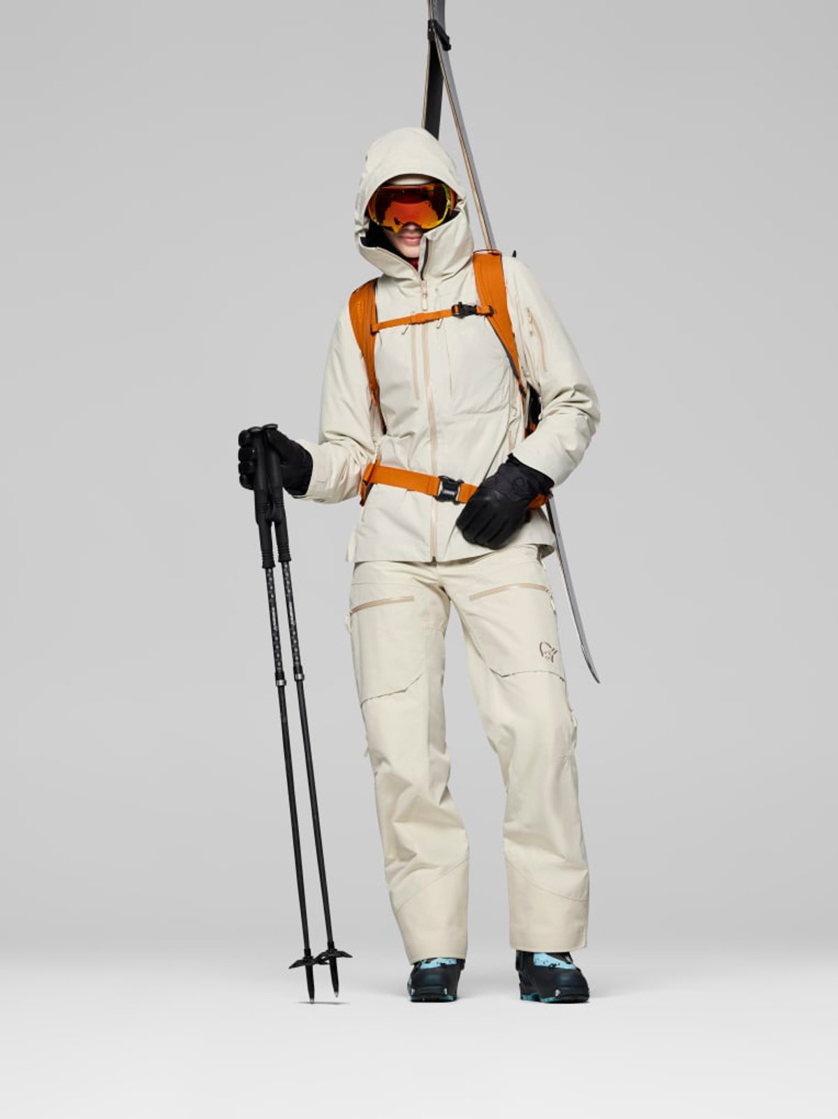 A skier wearing cream and white winter gear with orange accents, including a hooded jacket, snow pants, gloves, and orange ski goggles, holding ski poles with skis positioned over their shoulder against a white background.